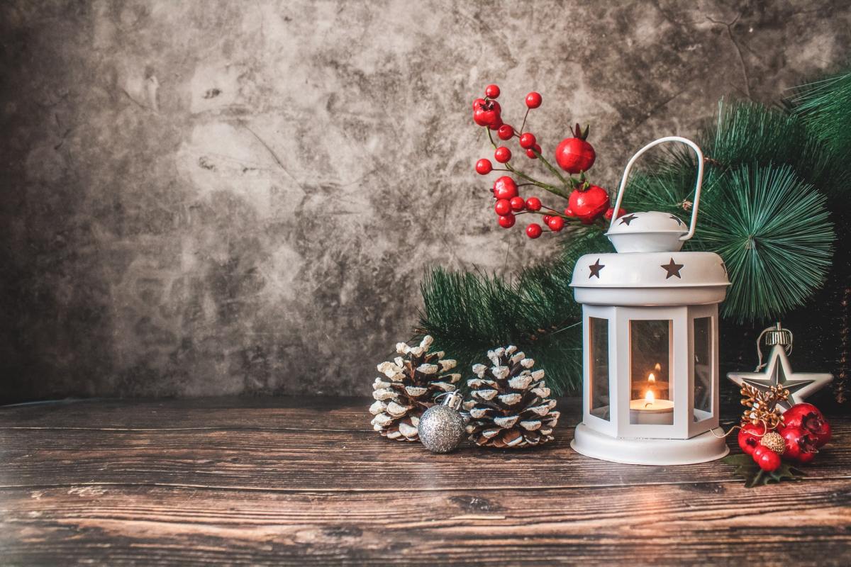 A tea light burns in a white lantern sitting next to a pine branch, pine cones, and red ornaments on a wood floor.